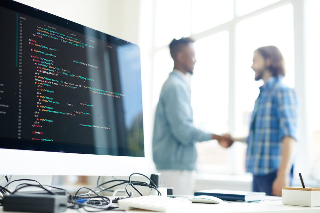 Zwei Kollegen schütteln sich im Hintergrund die Hände, im Vordergrund ist ein Monitor mit Code zu sehen.